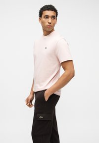 Homme portant un t-shirt rose à manches courtes et un pantalon noir avec des logos, debout avec une main dans la poche, regardant vers sa gauche sur un fond blanc.