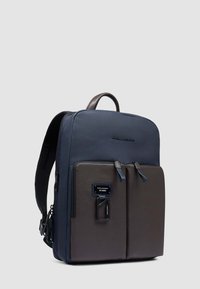 Mochila de cuero azul marino y marrón con diseño plano, dos bolsillos delanteros, correas para los hombros y un asa superior. Cuenta con una etiqueta con el logo y cremalleras.