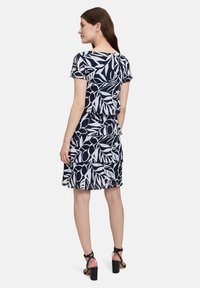 Granatowa sukienka z białym nadrukiem w liście, krótki rękaw, warstwowy projekt z falbanami i okrągłym dekoltem. Stylizowana z czarnymi sandałami na obcasie.