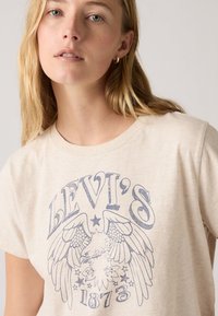 Lys beige bomull t-skjorte med blå grafikk av en ørn med vinger og teksten "LEVI'S 1873", rund hals og korte ermer.