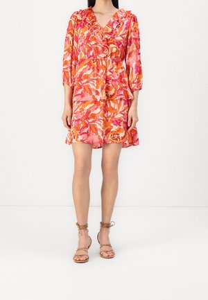 Kobieta ubrana w różowo-pomarańczową sukienkę kopertową w kwiaty z falbanami przy dekolcie w kształcie litery V oraz rękawami do połowy ramienia, w połączeniu z beżowymi sandałami na ramiączkach.