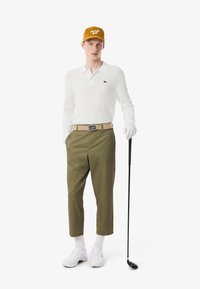 Polo a maniche lunghe bianco, pantaloni con motivo oliva, cintura beige, scarpe da golf bianche e cappello giallo con il testo "Lacoste Golf", mentre tiene una mazza da golf.
