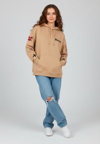 Felpa beige con tasca frontale, cappuccio con coulisse e dettagli ricamati. Abbinata a jeans blu strappati e sneakers bianche.