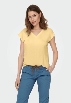 Vrouw draagt een geel T-shirt met korte mouwen en V-hals en een blauwe broek met een bruin gevlochten riem, staand met één hand in de zak.