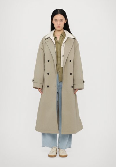 Light beige long coat with double-breasted buttons, wide collar, and textured fabric layered over a green shirt and blue jeans.