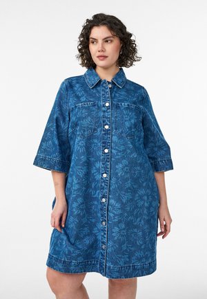 Kvinde med krøllet hår iført en midt-lang blå denimkjole med blomsterprint, frontknapper og brystlommer, stående foran en ensfarvet baggrund.