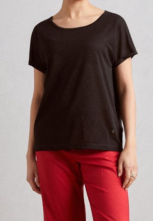 Person iført en løstsiddende sort kortærmet top med rund hals og knaldrøde bukser, stående med afslappede arme.