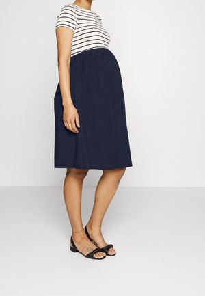 Kobieta w ciąży ubrana w krótki top w paski i granatową spódnicę do kolan oraz czarne sandały, stojąca bokiem.