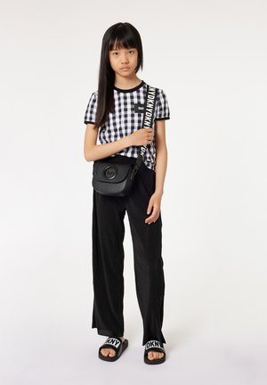 Sac à bandoulière noir avec une surface texturée, porté avec une chemise à carreaux noir et blanc, un pantalon large noir et des sandales à enfiler noires.