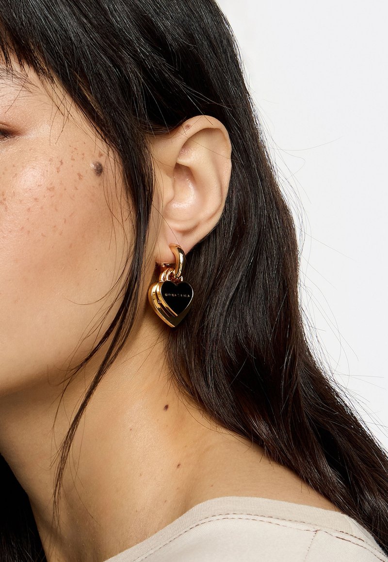 Pendientes en forma de corazón de oro con un centro negro brillante y un acabado dorado pulido. Textura detallada con un cierre de bisagra.