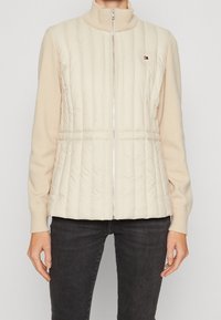 Beżowa pikowana kamizelka z prążkowanym kołnierzem i zamkiem błyskawicznym, o pikowanej fakturze i małym logo na górnej lewej stronie. Noszona na długich rękawach.