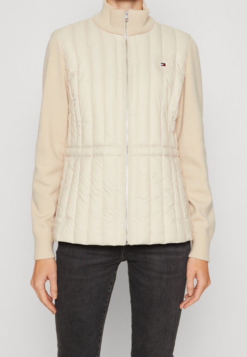 Beżowa pikowana kamizelka z prążkowanym kołnierzem i zamkiem błyskawicznym, o pikowanej fakturze i małym logo na górnej lewej stronie. Noszona na długich rękawach.