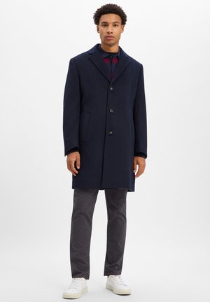 Homme debout portant un pardessus bleu marine foncé boutonné, un pantalon foncé, des baskets blanches et une chemise rayée sous le manteau.