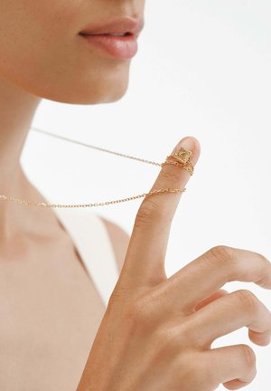 Anillo de cadena de oro se encuentra en la yema de un dedo, presentando un diseño cuadrado texturizado. La cadena se curva hacia atrás para colgar delicadamente. El tono de piel es neutro.