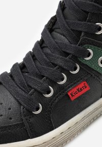 Zapatilla alta de cuero negro con cordones oscuros, ojales plateados, un acento texturizado verde y un pequeño logo de Kickers en rojo.