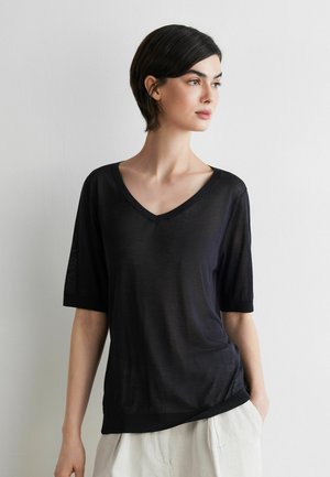 Frau mit kurzen dunklen Haaren, die ein schwarzes V-Ausschnitt-Kurzarmoberteil und helle Hose trägt und nach rechts blickt vor einem schlichten Hintergrund.