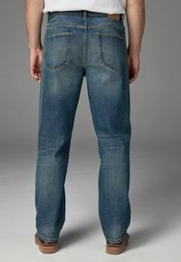 Jeans din denim albastru cu un design drept, având două buzunare pe spate și o etichetă pe talie, asortați cu cizme de lucru maro.