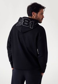 Felpa nera con grande grafica grigia "1981" sul retro, con una vestibilità rilassata, tessuto morbido e cappuccio con cordino.