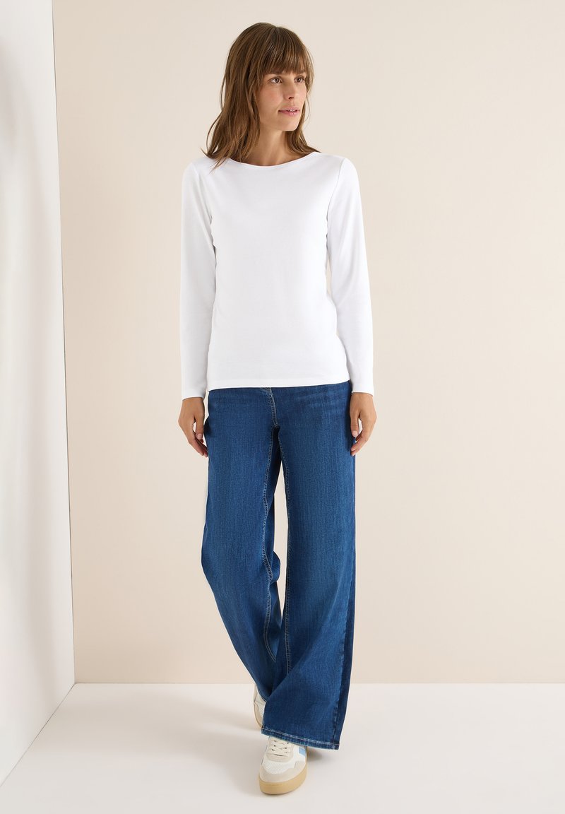 Weißes langärmliges Oberteil mit Rundhalsausschnitt, kombiniert mit weiten blauen Jeans und weißen Sneakers mit beigen Akzenten. Glatte Stoffoberfläche.