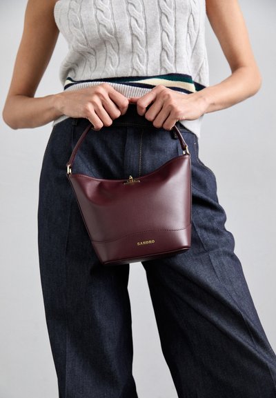 Borsa in pelle bordeaux con parte superiore curvata, manico corto, accessori in oro e logo impresso. Indossata con pantaloni ampi blu navy.