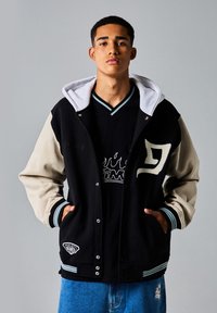 Zwarte varsity jacket met beige mouwen, grijze capuchon, geribbelde manchetten, voorste drukknoopsluiting en geborduurde logo details; gedragen over een zwart shirt.