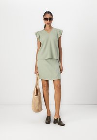 Top verde chiaro con maniche corte a volant e una gonna aderente abbinata. La modella tiene una borsa intrecciata, indossando occhiali da sole e scarpe col tacco lucide di colore scuro.