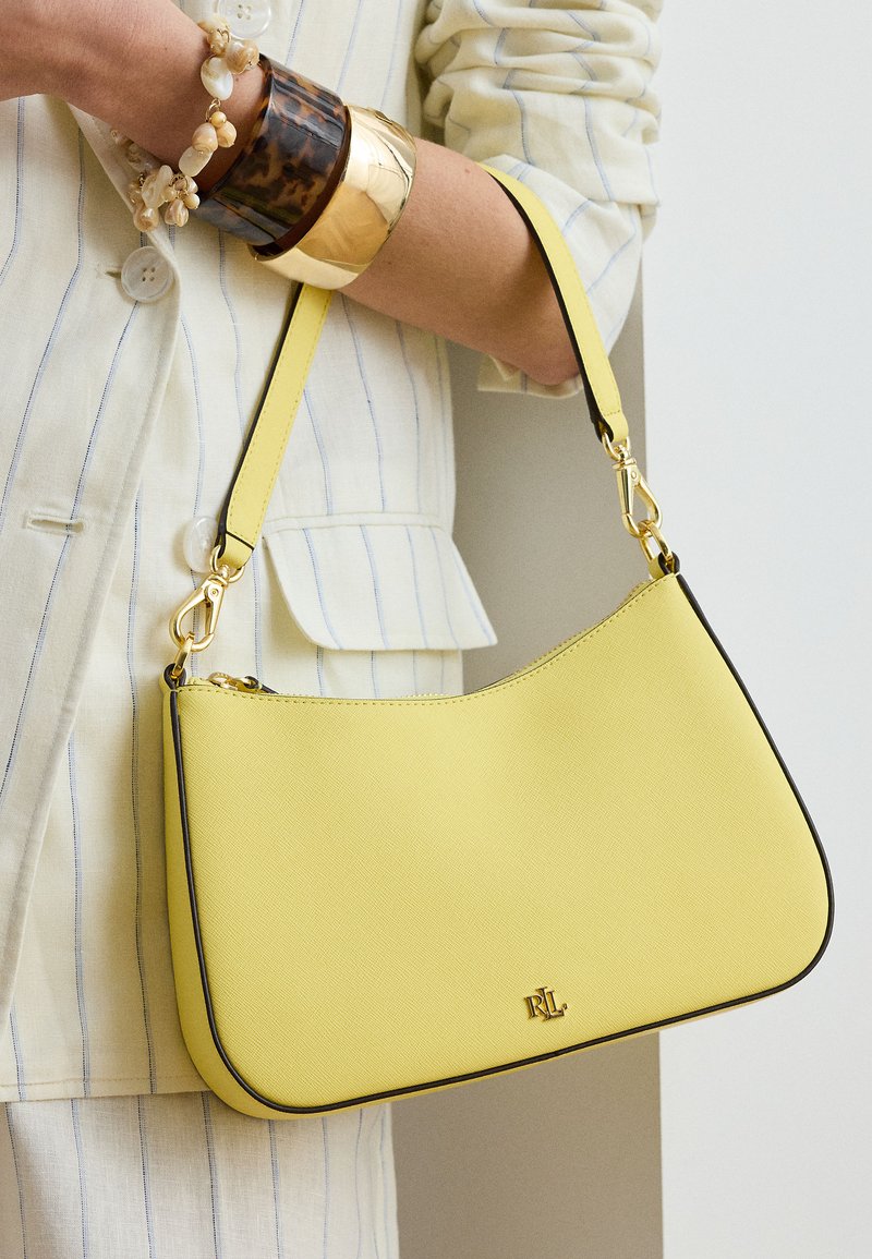 Κίτρινη δερμάτινη τσάντα με χρυσά μεταλλικά στοιχεία, κρατούμενη από άτομο που φοράει κοστούμι με εκρού ρίγες και στρωμένα βραχιολάκια στον καρπό.