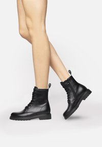 Bottines en cuir noir avec une texture lisse, devant à lacets et fermeture éclair sur le côté ; semelle épaisse et tirette à l'arrière pour un enfilage facile.