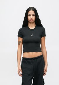 Γυναίκα με μακριά μαύρα μαλλιά, φορώντας μαύρο κοντό μπλουζάκι με άσπρο λογότυπο και μαύρο cargo παντελόνι, που στέκεται απέναντι από απλή φωτεινή background.