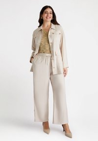 Beżowa kurtka z dużymi guzikami, noszona na tle fakturowanej złotej bluzki oraz luźnych beżowych spodni. Modelka ma na sobie beżowe szpilki.