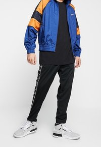 Blå och orange träningsjacka med dragkedja fram, svart t-shirt, svarta byxor med vit sidostrip, och grå sneakers med vita snörning.