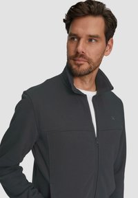 Chaqueta gris con cremallera y cuello alto, de tejido texturizado, con una costura cosida en el pecho. Presenta un logo sutil en el lado izquierdo.