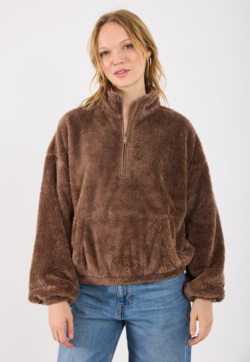 Sudadera de forro polar marrón con cuello con cremallera, corte holgado y mangas anchas. Presenta un tejido texturizado y suave, además de un bolsillo tipo canguro en la parte frontal.
