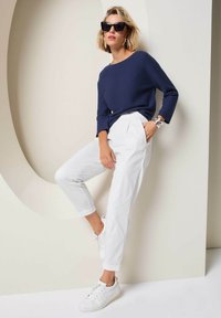 Marineblauer Pullover, weiße schmal geschnittene Hose und weiße Sneakers. Das Modell trägt übergroße schwarze Sonnenbrillen; der Hintergrund zeigt sanfte neutrale Töne.