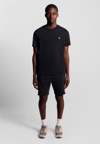 Svart t-shirt med liten gul logotyp, ihop med svarta shorts. Modell som bär ljusa sneakers och vita strumpor, står mot en grå bakgrund.