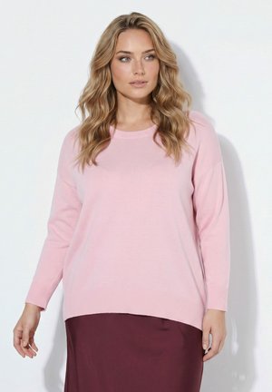 Kvinde med langt bølget blondt hår iført en lyserød sweater og et mørkerødt skørt, der står foran en hvid baggrund.