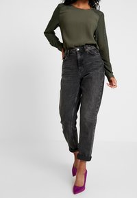Donna che indossa una blusa verde oliva a maniche lunghe, jeans neri a vita alta con risvolti e scarpe con tacco viola, in piedi su uno sfondo bianco.