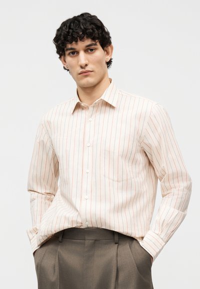 Νεαρός άνδρας με σγουρά σκούρα μαλλιά, φοράει μπεζ πουκάμισο με λεπτές κόκκινες κάθετες ρίγες και καφέ παντελόνι, τα χέρια στις τσέπες, ουδέτερο φόντο.