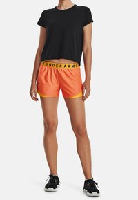 Svart kort t-shirt, orange sportshorts med gul midjeresår och svarta sneakers. Mjukt tyg, avslappnad passform, kontrasterande färger.