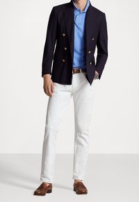 Marine dubbelzijdige blazer, lichtblauw polo, witte broeken en bruine instappers. Opvallende goudkleurige knopen op de blazer. Glad, op maat gemaakte pasvorm.