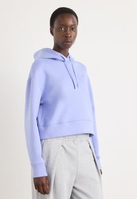 Hoodie lavande court avec cordon de serrage et logo brodé, associé à un pantalon de jogging plissé gris clair. Tissu doux avec une texture lisse.