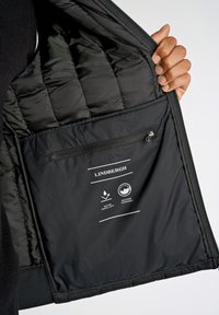 Svart puffjacka inuti med quiltat foder, dragkedjeficka och skötselråd som visar vattenavvisande och maskintvättbara ikoner.