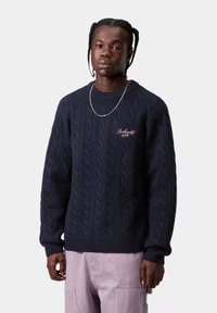 Granatowy sweter z dzianiny w warkocze z prążkowanymi mankietami i dolnym brzegiem, z subtelnym haftem logo na piersi. Noszony z jasnofioletowymi spodniami cargo.