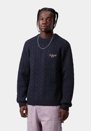 Suéter de punto grueso azul marino con puños y dobladillo acanalados, con un bordado sutil del logo en el pecho. Combinado con pantalones cargo de color púrpura claro.