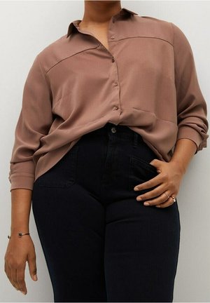 Blouse marron à manches longues avec une fermeture à boutons sur le devant, présentant deux coutures horizontales traversant la poitrine. Associée à un jean denim foncé.