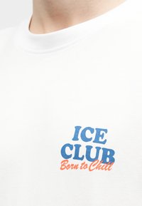 Λευκό μπλουζάκι με στρογγυλή λαιμόκοψη, με μπλε κείμενο "ICE CLUB" και πορτοκαλί καλλιγραφικό κείμενο "Born to Chill" στο στήθος.