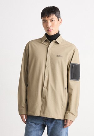 Beige long-sleeve shirt with button front and black patch pocket; features a collar, smooth fabric, and side zip pockets.
