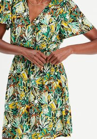 Robe florale avec décolleté en V, manches courtes et taille élastique. Les couleurs prédominantes comprennent le vert, le jaune et le brun avec des motifs élaborés.