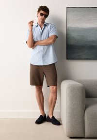 Camicia a righe color azzurro chiaro a maniche corte, pantaloncini marroni e mocassini blu navy. La persona è in piedi accanto a un divano grigio e a un dipinto astratto.