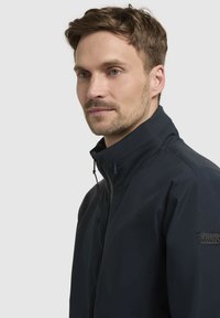 Tengerészeti vízálló dzseki magas gallérral, cipzárral és állítható húzózsinórral. Az ujjon márkázott textil patkó található.